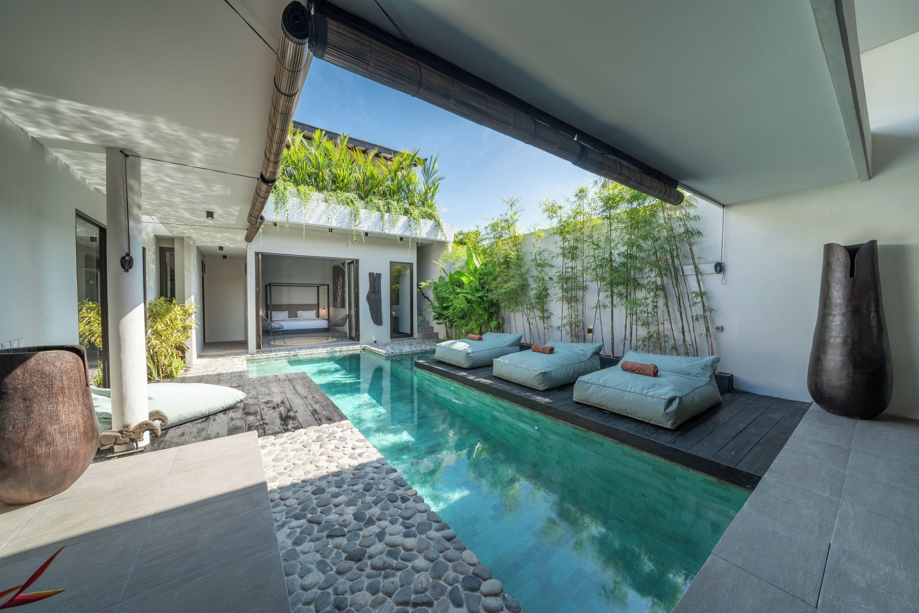A modern outdoor pool area with a stone pathway, seating with cushions, and a bamboo garden.