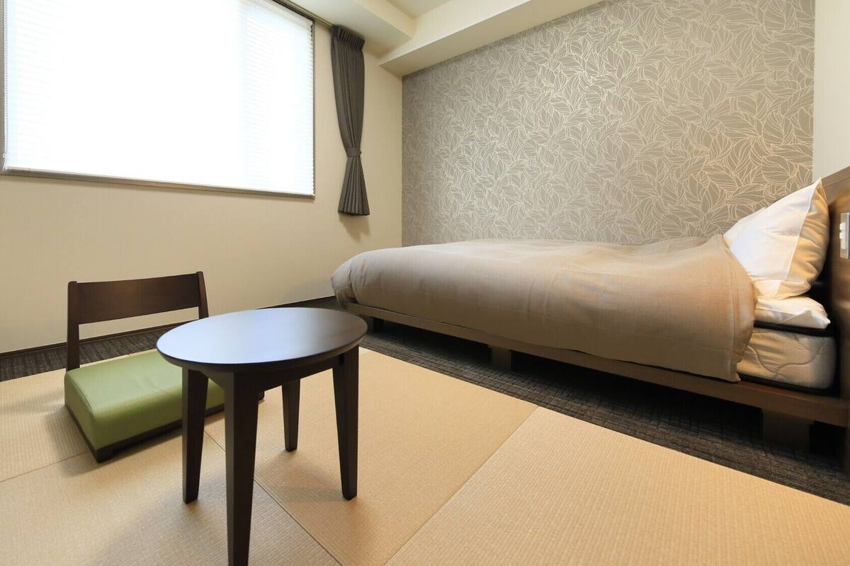 Ein Hotelzimmer mit Bett, Stuhl, kleinem Tisch und einem Fenster mit Vorhängen.