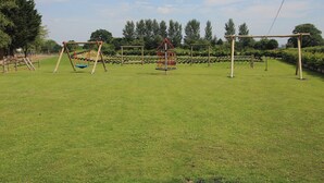 A playground with swings and climbing structures, surrounded by a grassy field and trees. - Swan (Leominster)