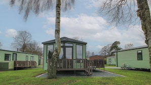 A green mobile home with a fenced deck, surrounded by trees and other similar homes.