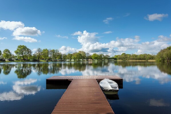 Un lago tranquilo con un muelle de madera que se adentra en el agua, rodeado de árboles y cielos despejados.
