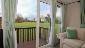 A room with a sliding glass door leading to a deck, a beige sofa with a green pillow, and a view of a grassy area with trees. - Owl (Leominster)