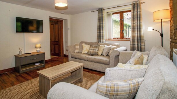A living room with a sofa, a coffee table, a TV mounted on the wall, a lamp, and a window with curtains. - Stables Cottage (Leominster)