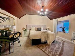 A bedroom with a large bed, a chandelier, a window with a view, and a desk with a chair. - Style House Luxury Cottage (Jameston)