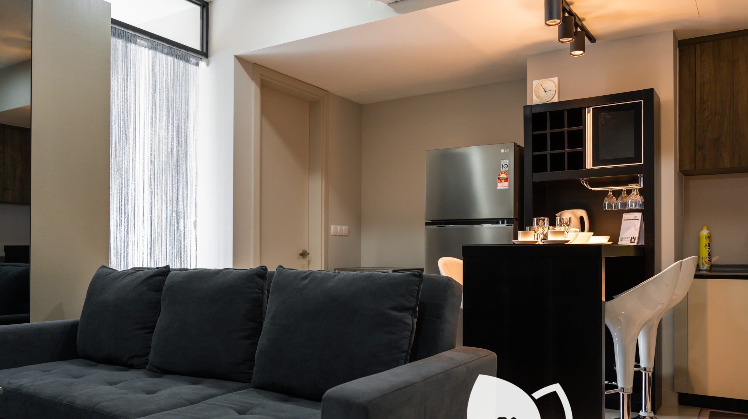 A modern living room with a dark grey sofa, a kitchen area with a refrigerator and a small bar counter, and a window with sheer curtains.