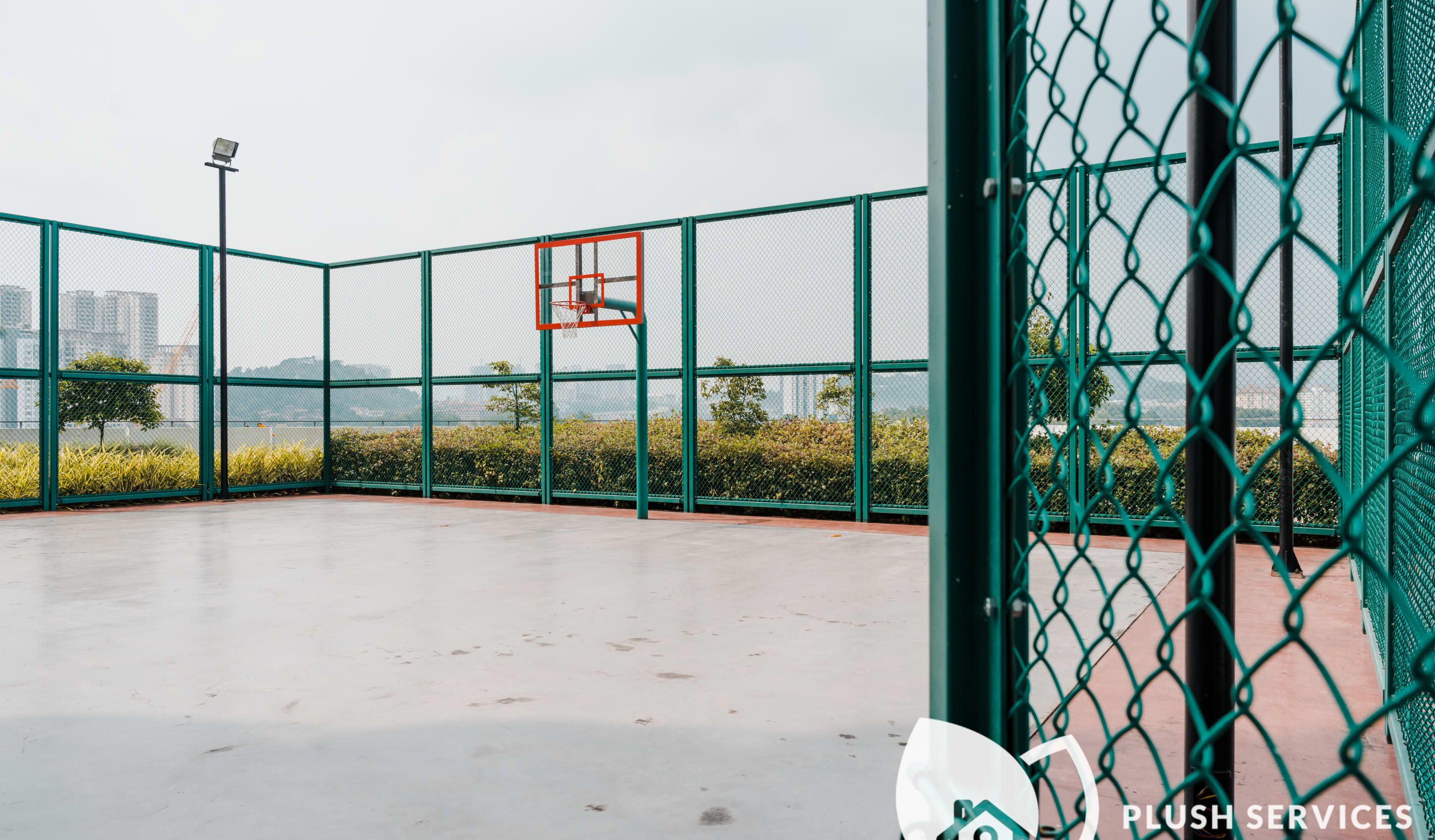 An outdoor basketball court with a green fence, a basketball hoop, and a view of buildings in the background.
