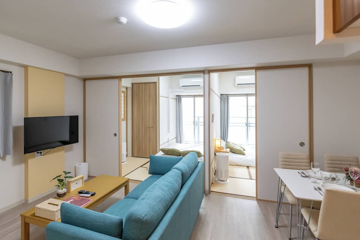 Un salon moderne avec un canapé bleu, un téléviseur à écran plat, une table basse en bois et un coin repas comprenant une table et des chaises blanches.