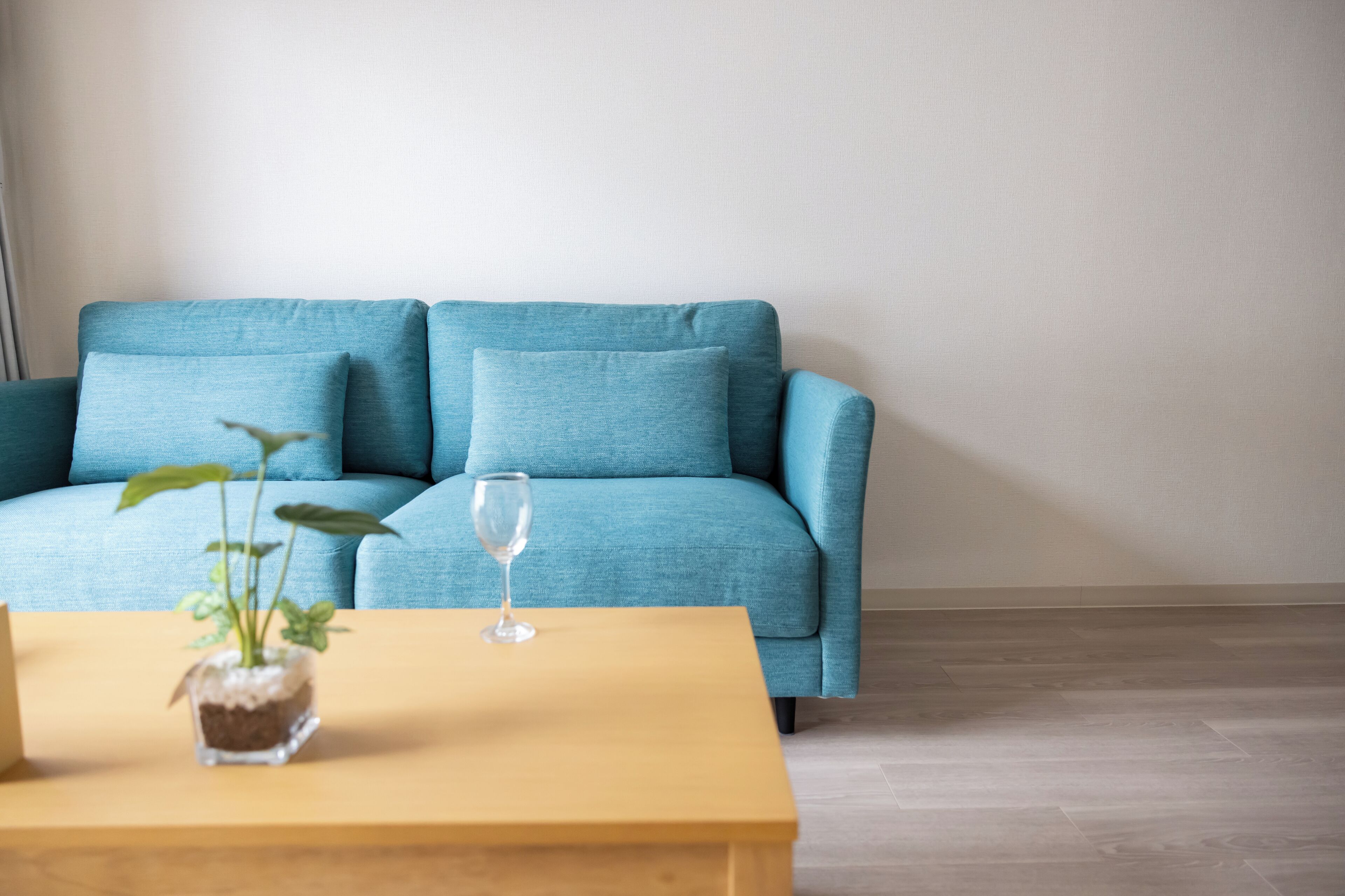 A blue sofa with two cushions, a small potted plant, and a clear glass on a yellow table in a room with a plain wall and wooden flooring.