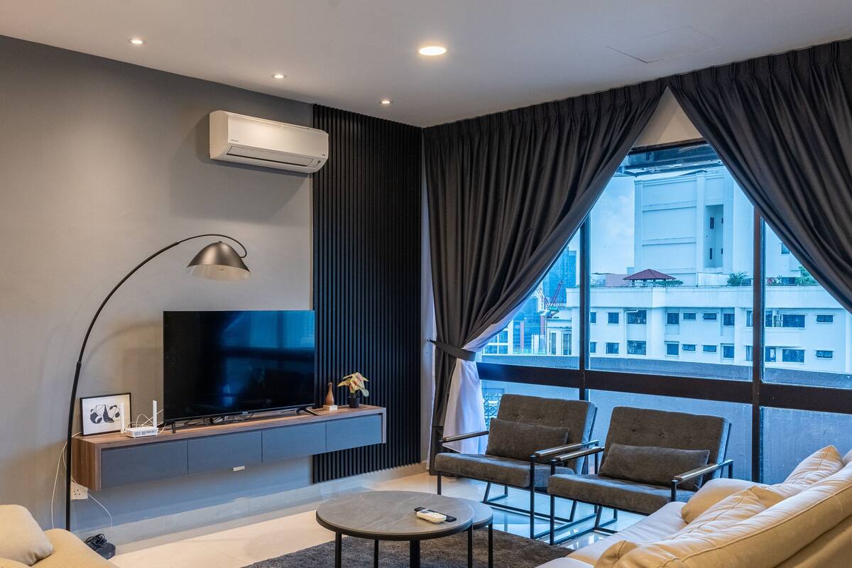 Een moderne woonkamer met een flatscreen televisie, comfortabele zitmogelijkheden, een groot raam met gordijnen, en een airconditioning.