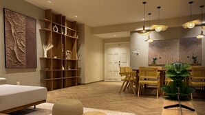 Un salon moderne comprenant un canapé, un plancher en bois, des poufs en fibres naturelles, un coin repas avec des chaises et une table, ainsi qu’une grande œuvre d’art murale.