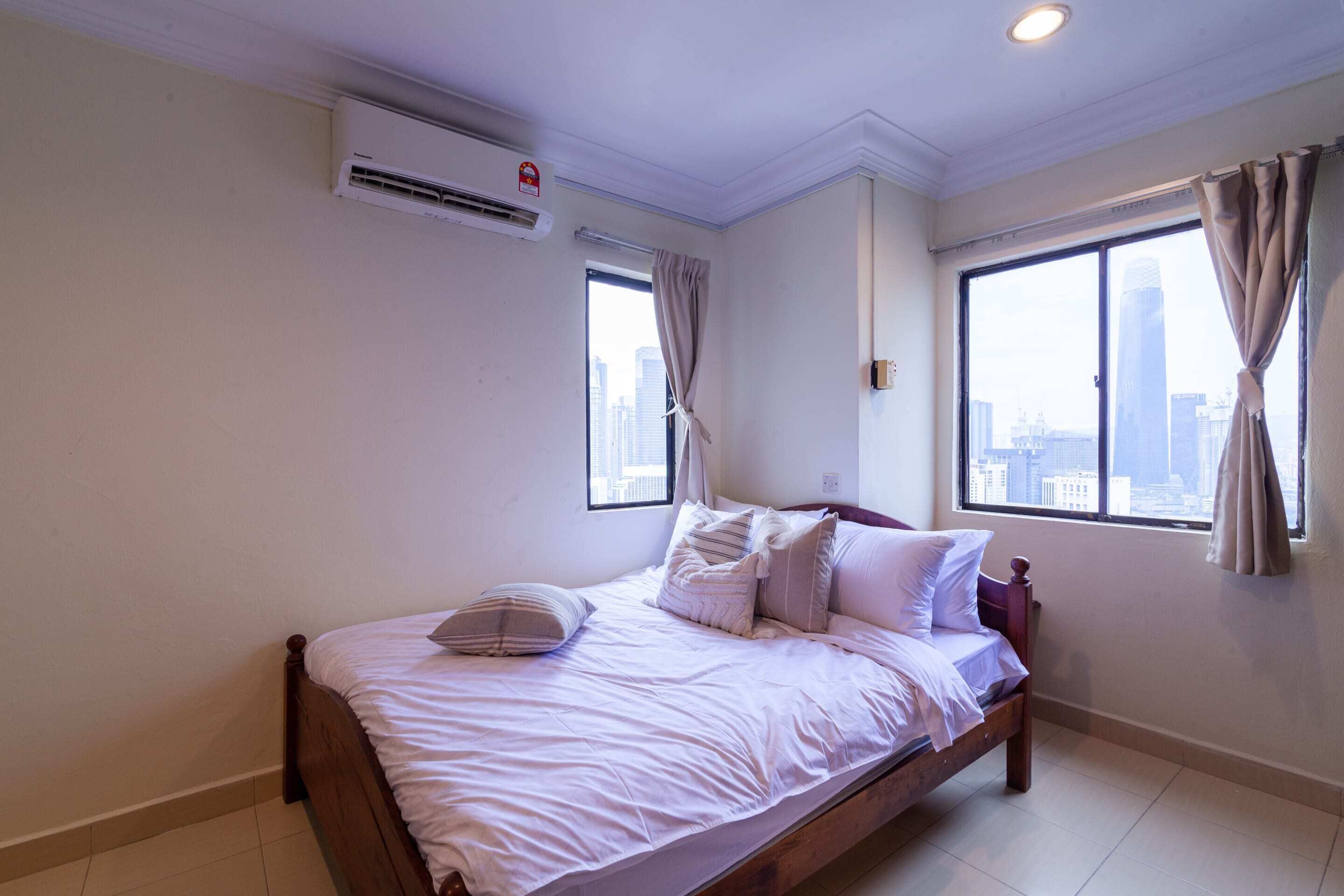 Un dormitorio con cama, aire acondicionado, dos ventanas con cortinas y vistas a la ciudad.