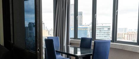 A hotel room with a large window, a glass table, and two blue chairs.