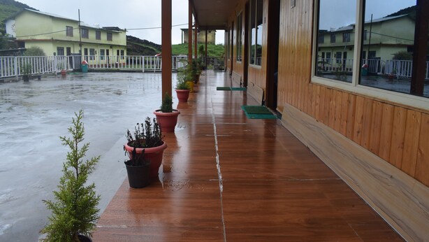 A covered outdoor area with a wooden floor, potted plants, and a view of buildings in the background. - Chopta Himrab Resort  (Pokhri)