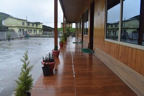 A covered outdoor area with a wooden floor, potted plants, and a view of buildings in the background. - Chopta Himrab Resort  (Pokhri)