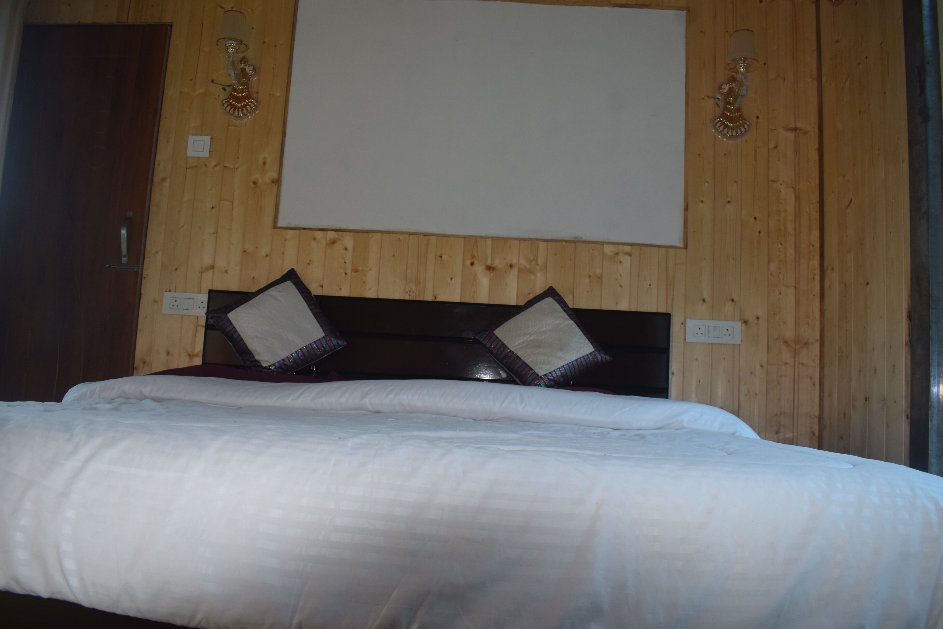 A hotel room with a wooden wall, a bed with white bedding, and two decorative wall sconces.