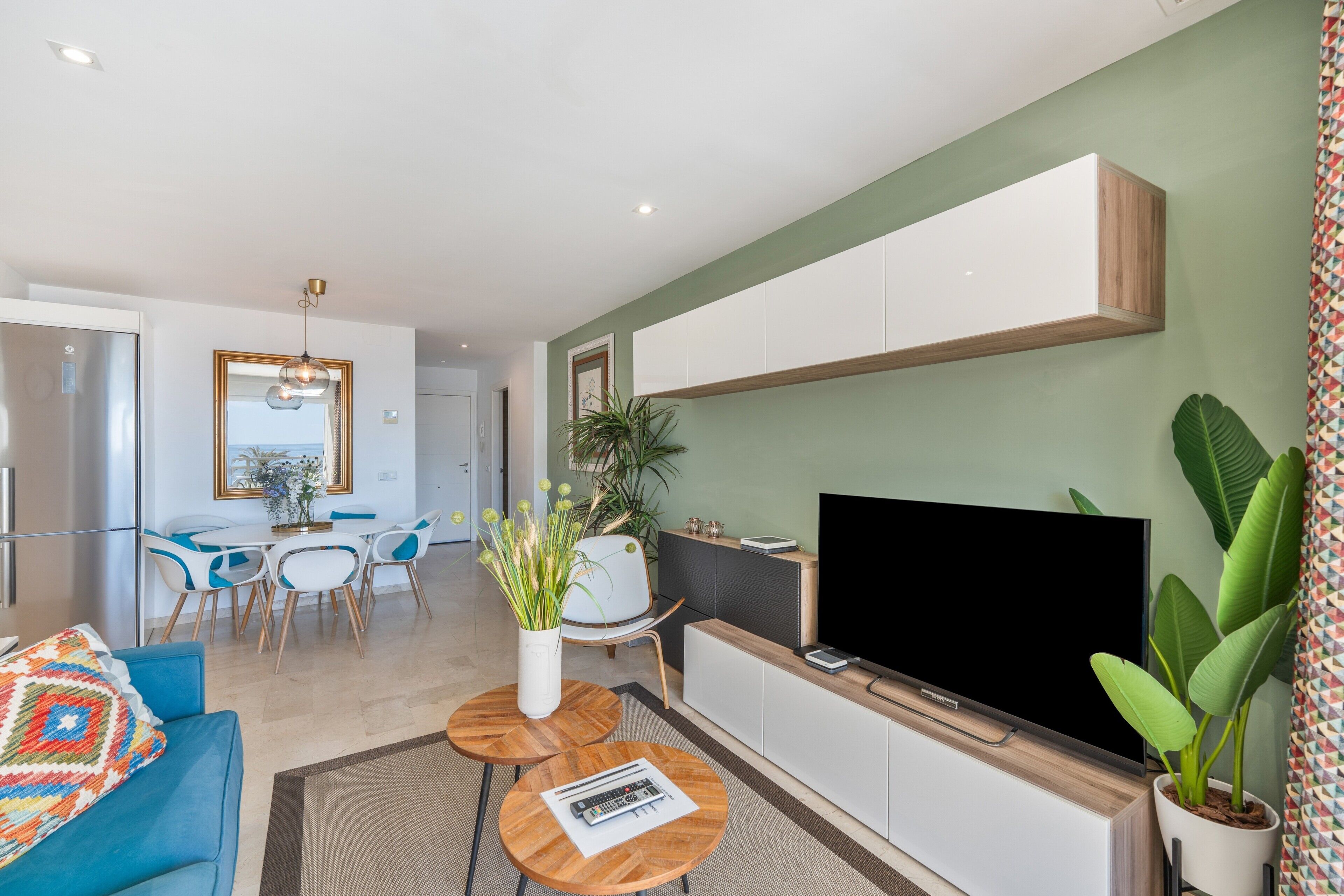 A modern living room with a flat-screen TV, a blue sofa, a dining area with a white table and chairs, and a large plant.