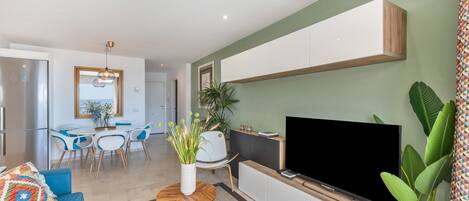 A modern living room with a flat-screen TV, a blue sofa, a dining area with a white table and chairs, and a large plant.