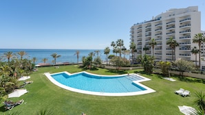 A swimming pool surrounded by grass and palm trees, with a view of the sea and a multi-story building in the background. - Front Line Beach 2-Bed Apartment with Sea Views, Wi-Fi, and Air Conditioning (Marbella)