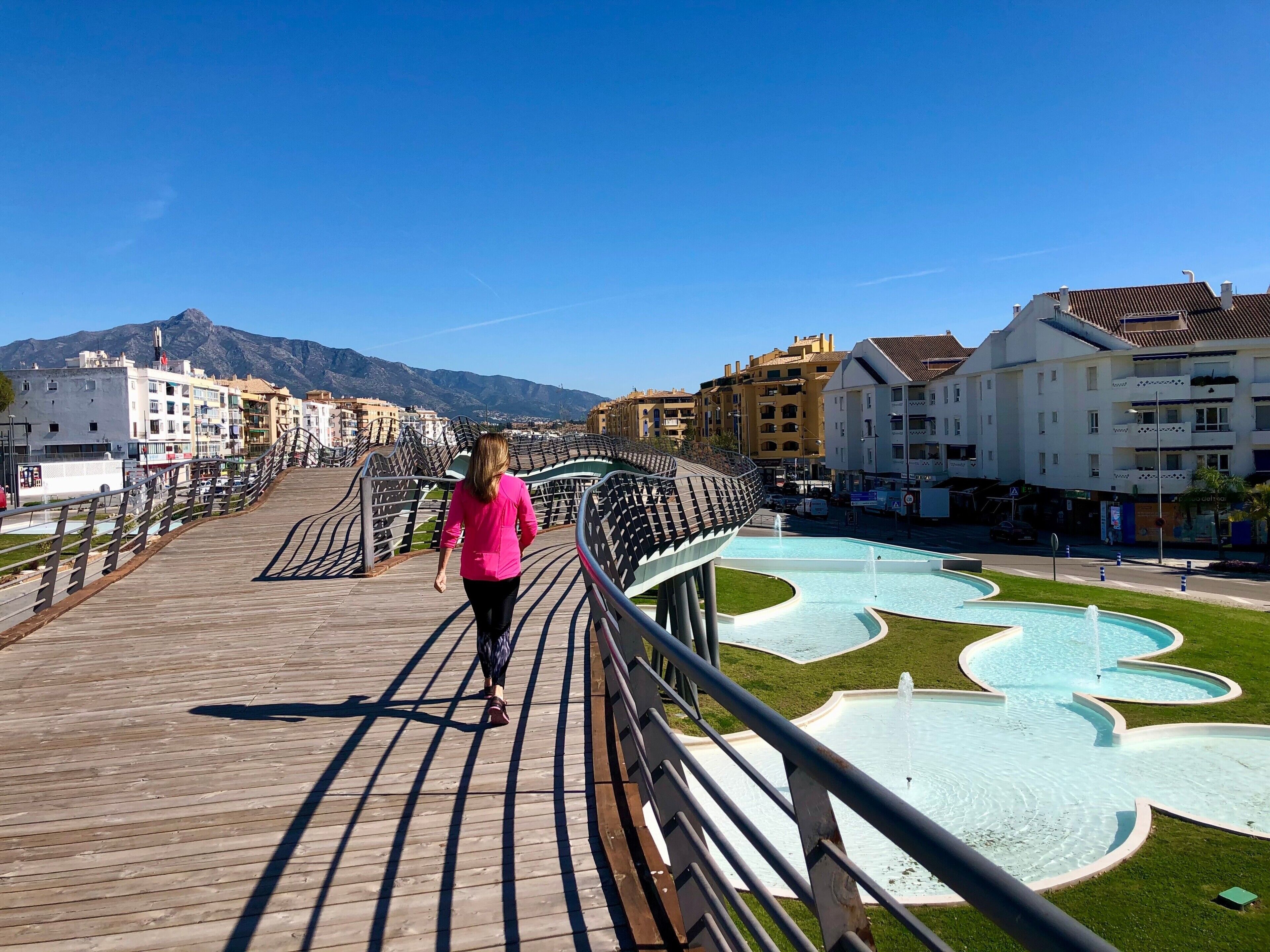 Un ponte pedonale con un design curvo particolare, un'area con fontana e edifici sullo sfondo.