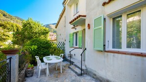 A stone-floored patio with a table and chairs, a white wall with green shutters, and a mountain backdrop. - Holiday Home 'Jolie Maison' with Wi-Fi (Lucéram)