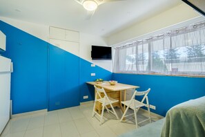 Una habitación con pared azul, una mesa de madera con dos sillas, un televisor montado en la pared y una ventana con cortina.