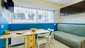 Ein Zimmer mit Esstisch, einer Couch, einem Fenster mit Vorhang und einem an der Wand montierten Fernseher.