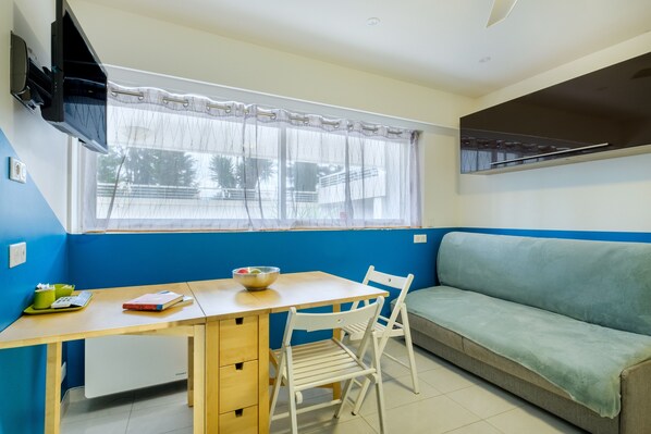 Una habitación con una mesa de comedor, un sofá, una ventana con cortina y un televisor montado en la pared.