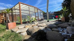 A glass greenhouse structure with chairs inside, surrounded by rocks and plants.