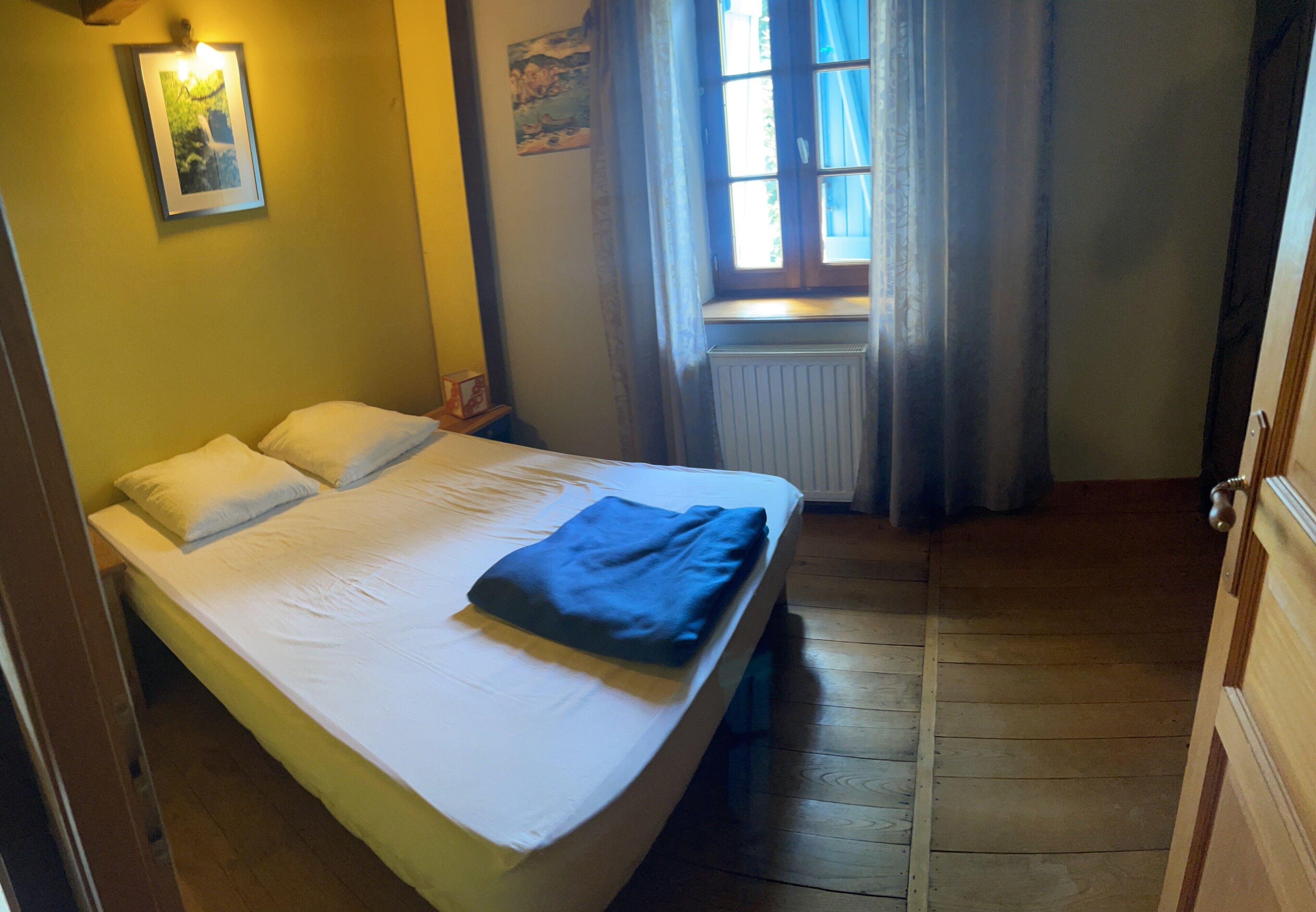 Un dormitorio con cama, ventana con cortinas, radiador y un cuadro en la pared.