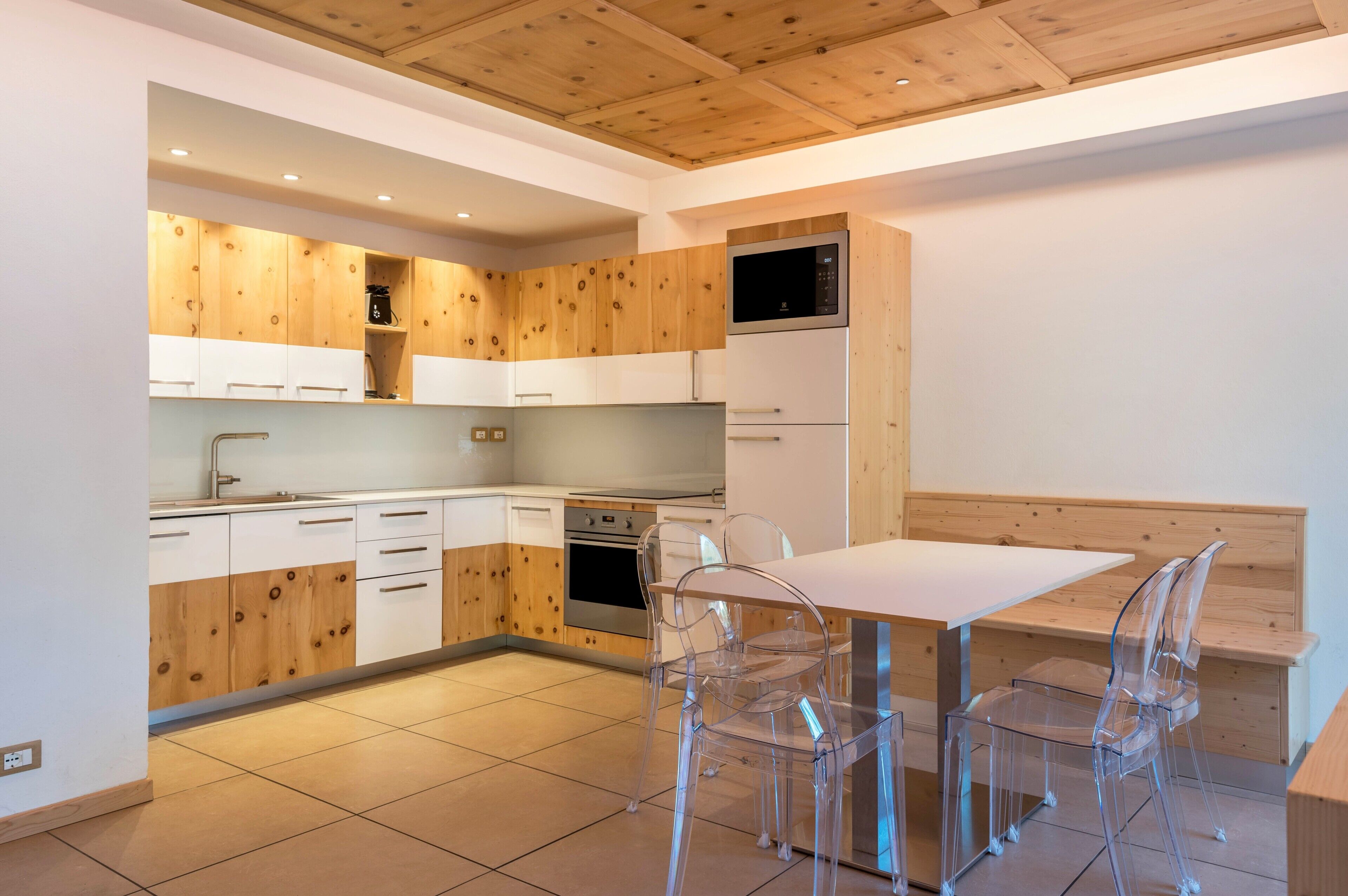 Une cuisine moderne avec des meubles en bois, un four à micro-ondes encastré et un coin repas comprenant une table blanche et des chaises transparentes.