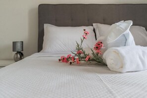 Un lit bien fait, avec du linge de lit blanc, une tête de lit grise et une petite table de chevet sur laquelle se trouve une lampe.