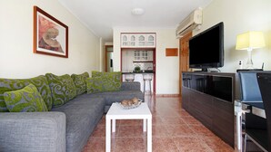 Un salón con un sofá gris, una mesa de centro blanca, un televisor y una zona de cocina al fondo.