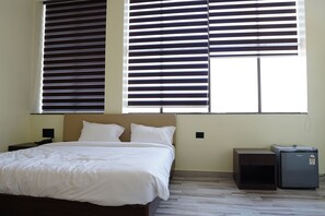 A bedroom with a bed, two bedside tables, a nightstand, a small refrigerator, and large windows with blinds.