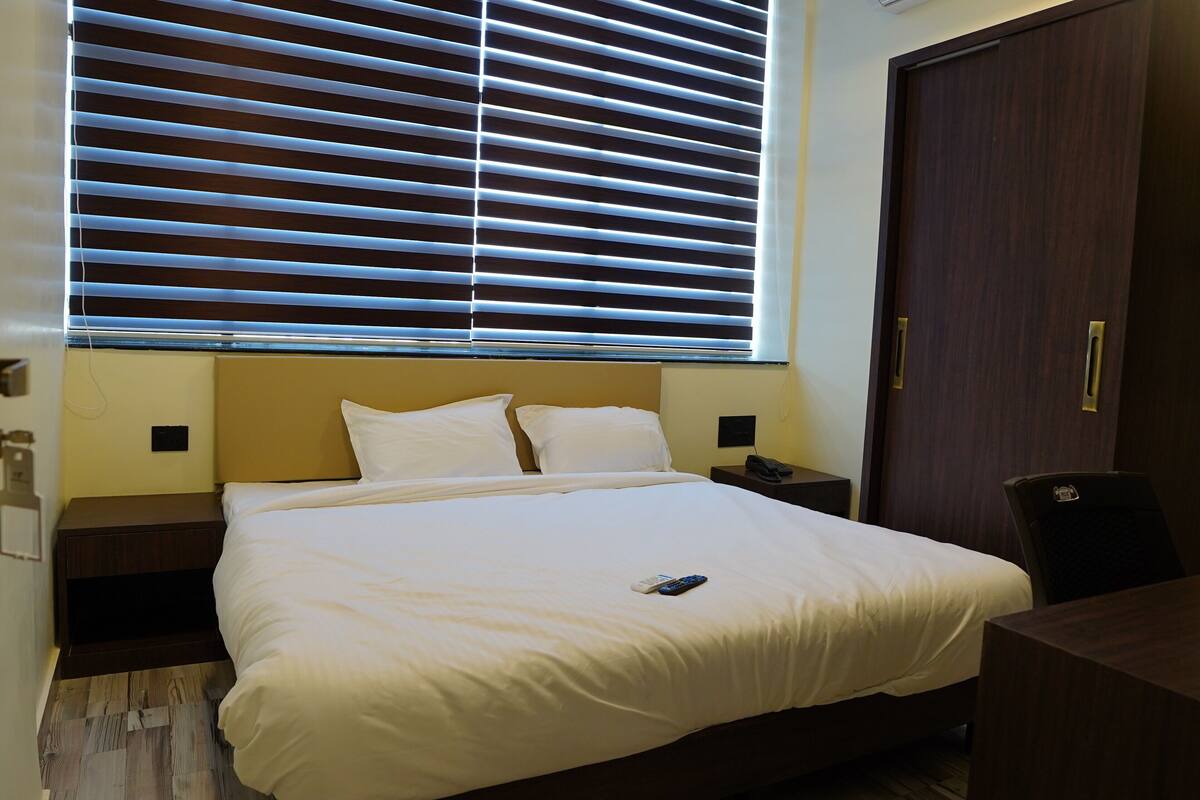 Uma cama bem arrumada com lençóis brancos, uma mesa de cabeceira com abajur e um guarda-roupa de madeira.
