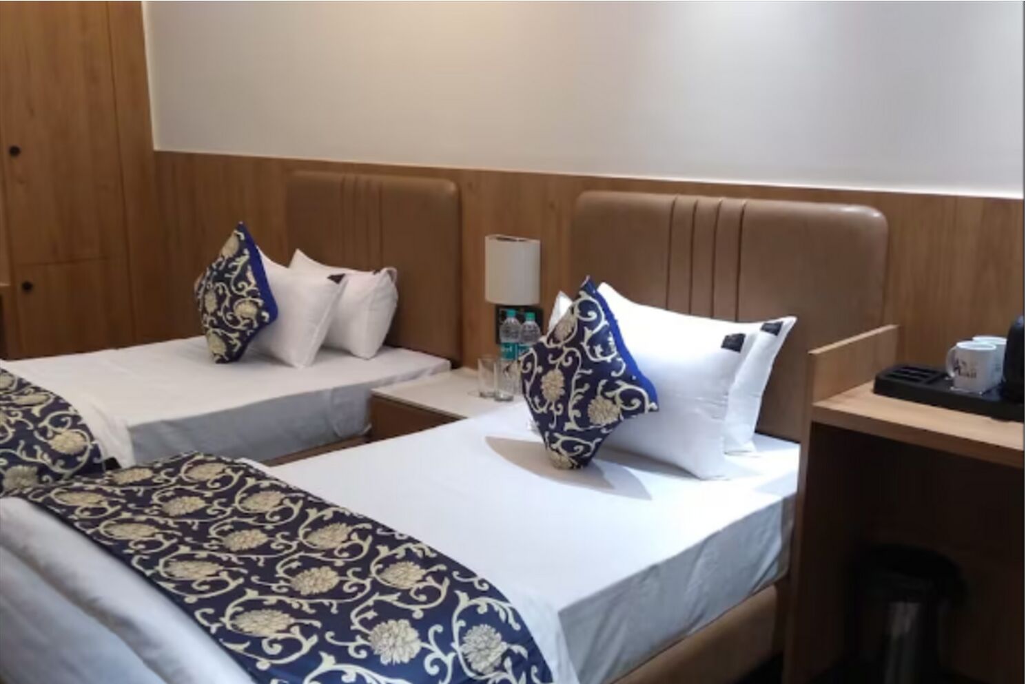 Une chambre d’hôtel avec deux lits, chacun recouvert d’une couvre-lit à motifs et agrémenté d’oreillers assortis. On y trouve une table de chevet avec une lampe et une bouteille d’eau.