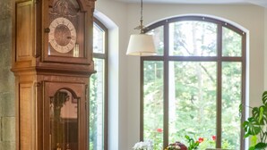 A dining area with a wooden clock, a vase with flowers, and a view of greenery through large windows. - Opus Villa (Sinaia)