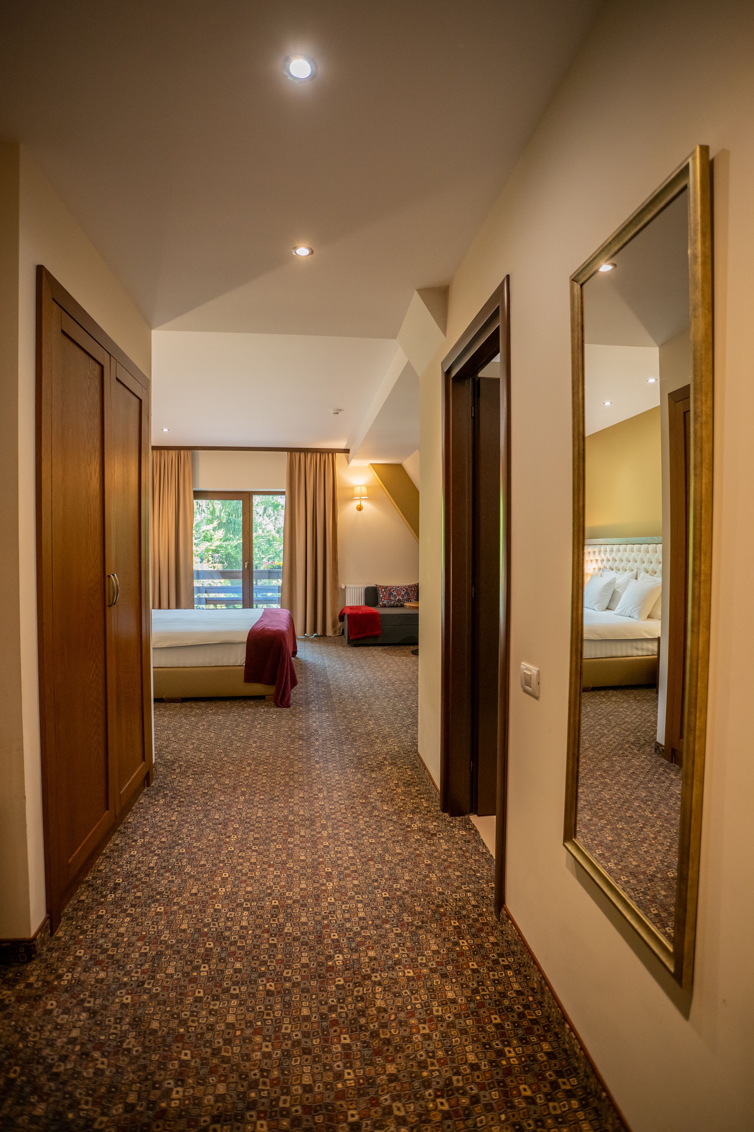 A hotel hallway with a patterned carpet, a bed with a red blanket, and a large mirror on the wall.