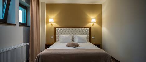 A hotel room with a large bed, two wall-mounted lamps, a radiator, and a window with curtains.