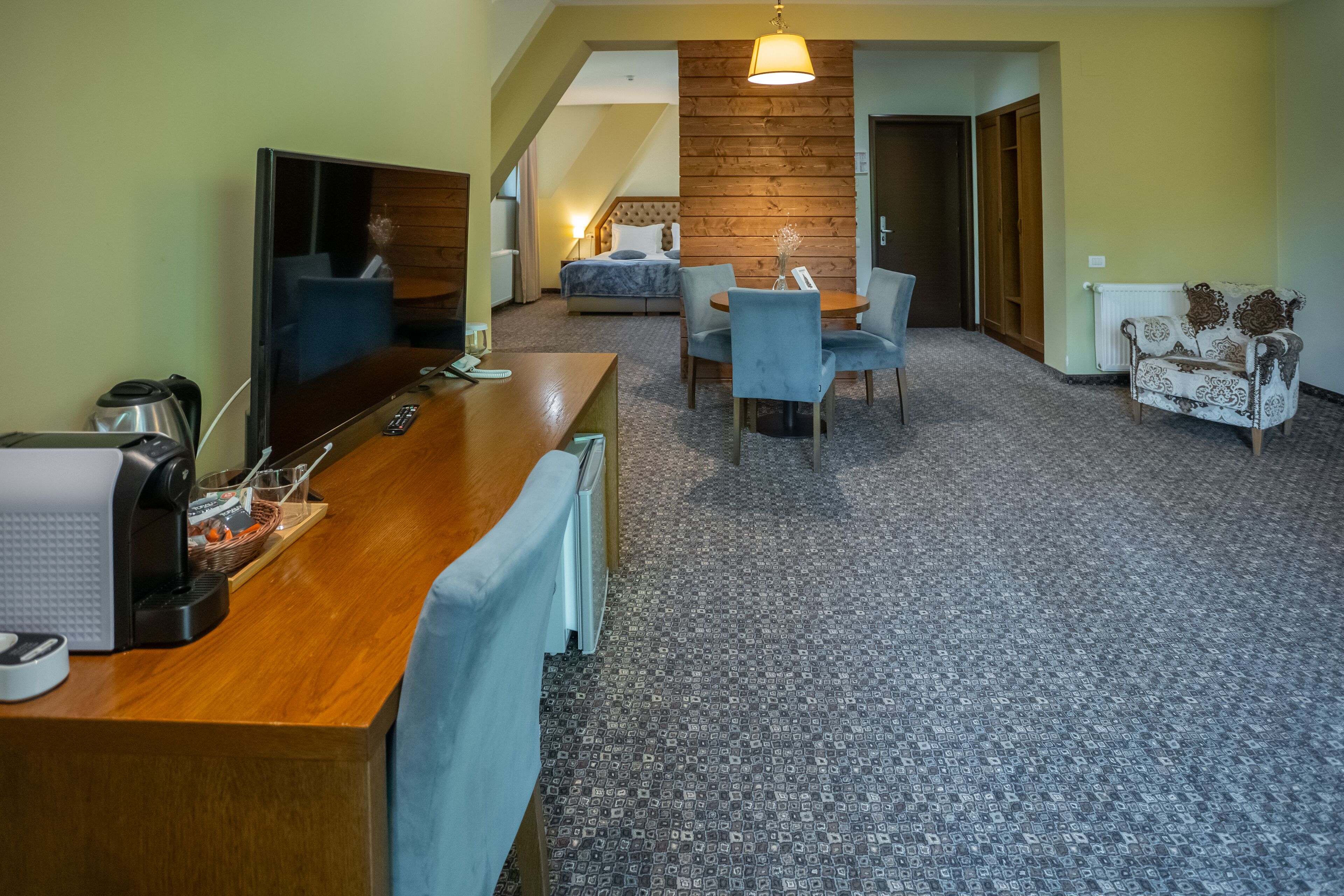 A hotel room with a wooden desk, a television, a bed, a dining area with chairs, and a patterned carpet.