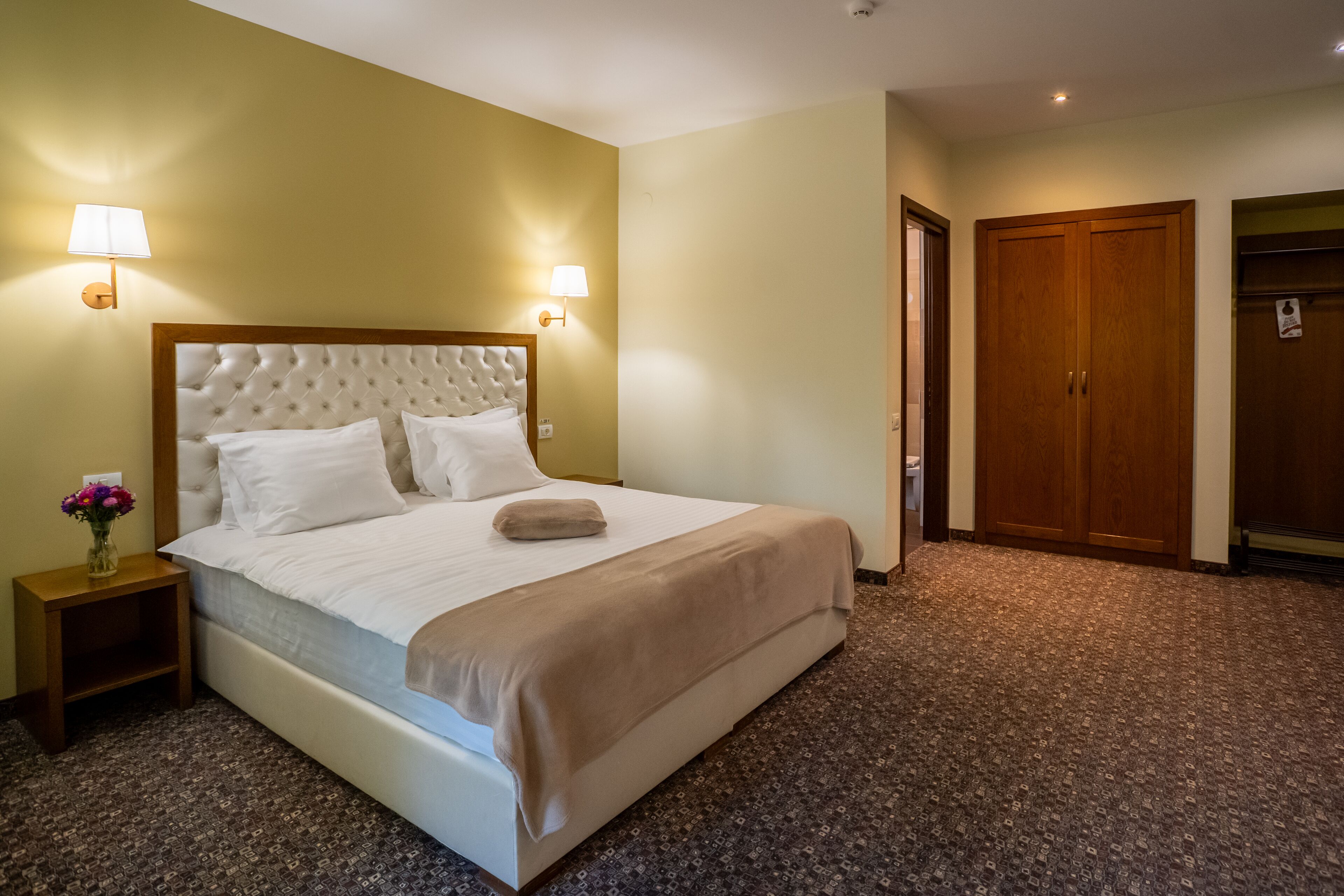 A hotel room with a large bed, two bedside lamps, a wooden wardrobe, and a carpeted floor.