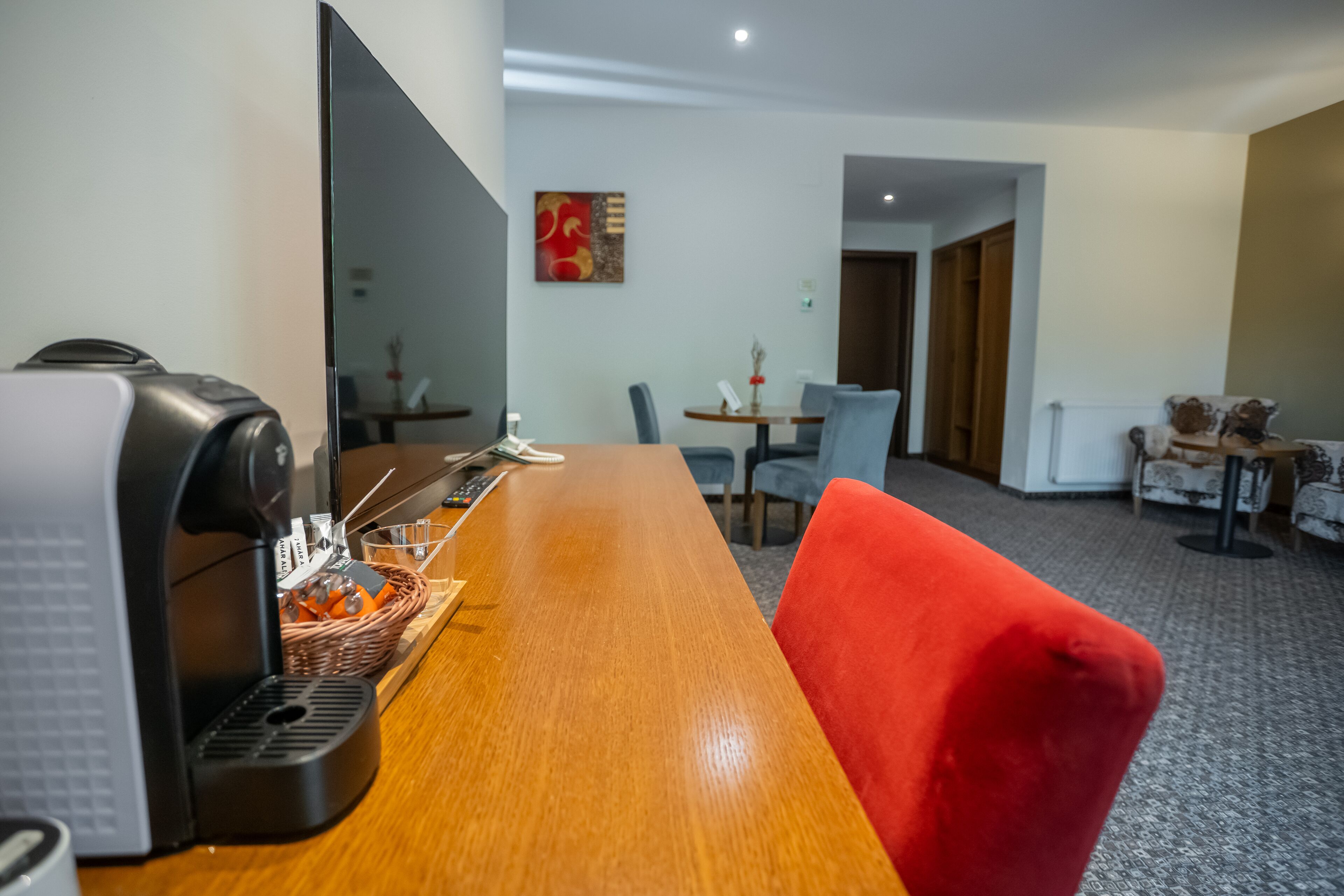 Een hotelkamer met een houten bureau, een rode stoel, een koffieapparaat en een zithoek met een tafel en stoelen.
