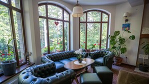 A cozy room with a curved black leather sofa, a wooden coffee table, and a green armchair. Large windows with a view of trees and flowers are visible. - Opus Villa (Sinaia)