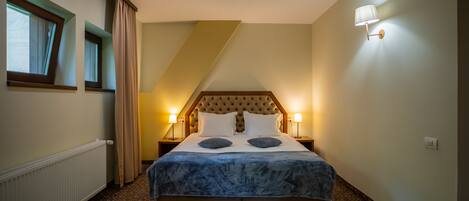 A hotel room with a large bed, two bedside lamps, a radiator, and a window with curtains.