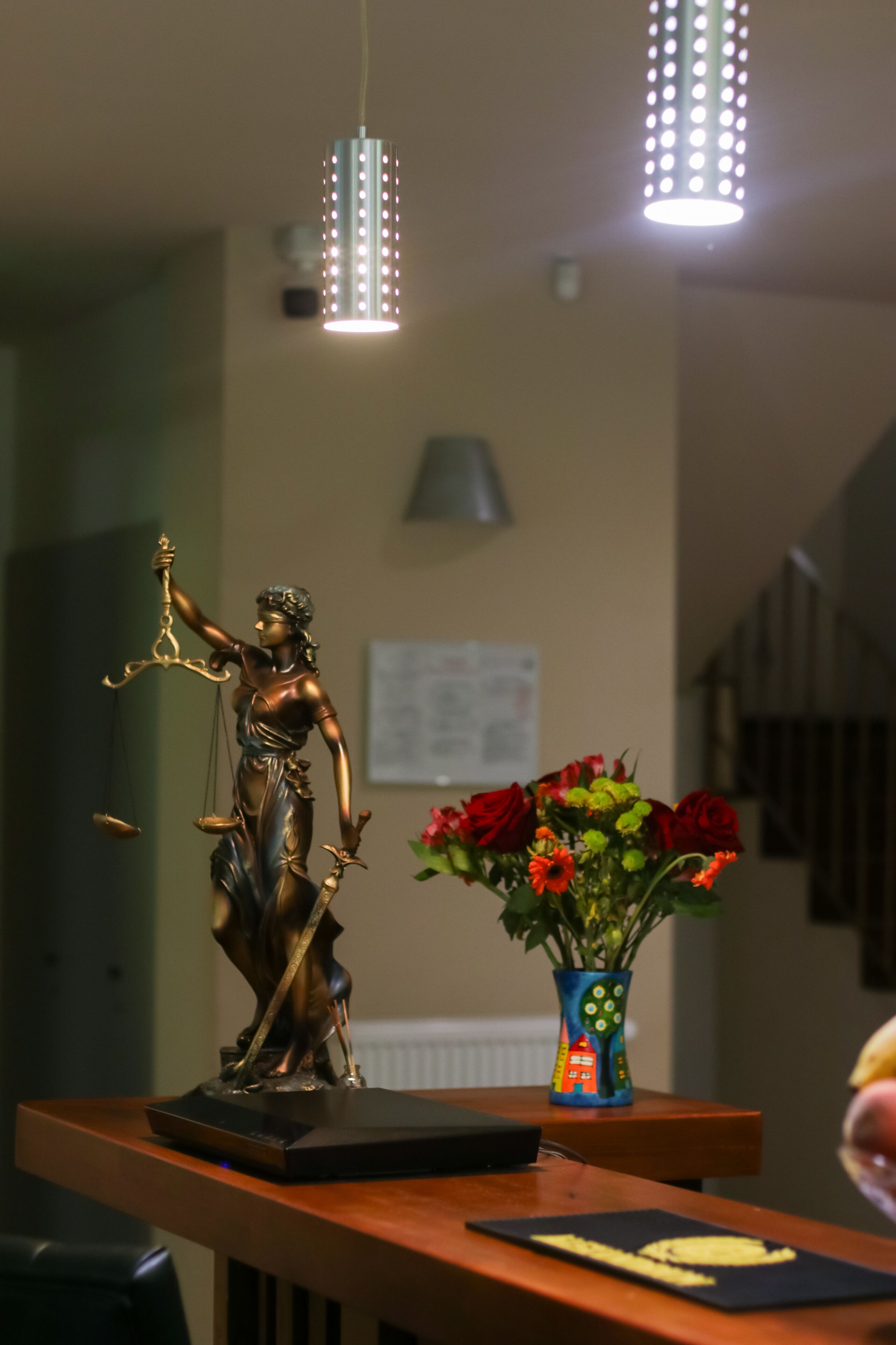 Una estatua de la Justicia sobre una mesa con un jarrón de flores y una lámpara al fondo.