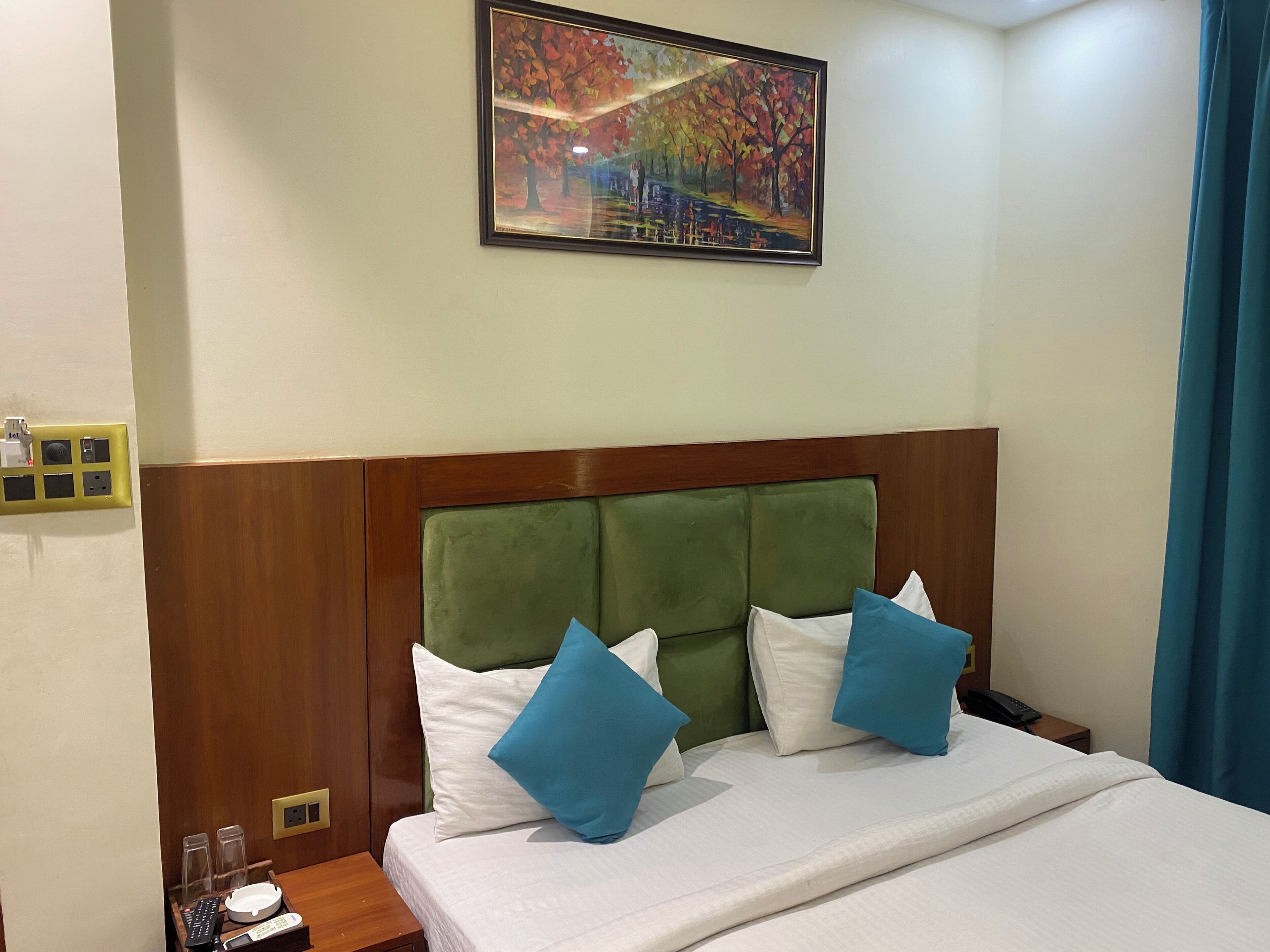 A hotel room with a bed, a headboard, a painting, and a nightstand with a phone and glasses.