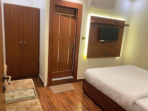 A bedroom with a wooden wardrobe, a door with a handle, a bed with a white mattress, a small rug, and a wall-mounted TV. - Ramesh Palace (Prayagraj)