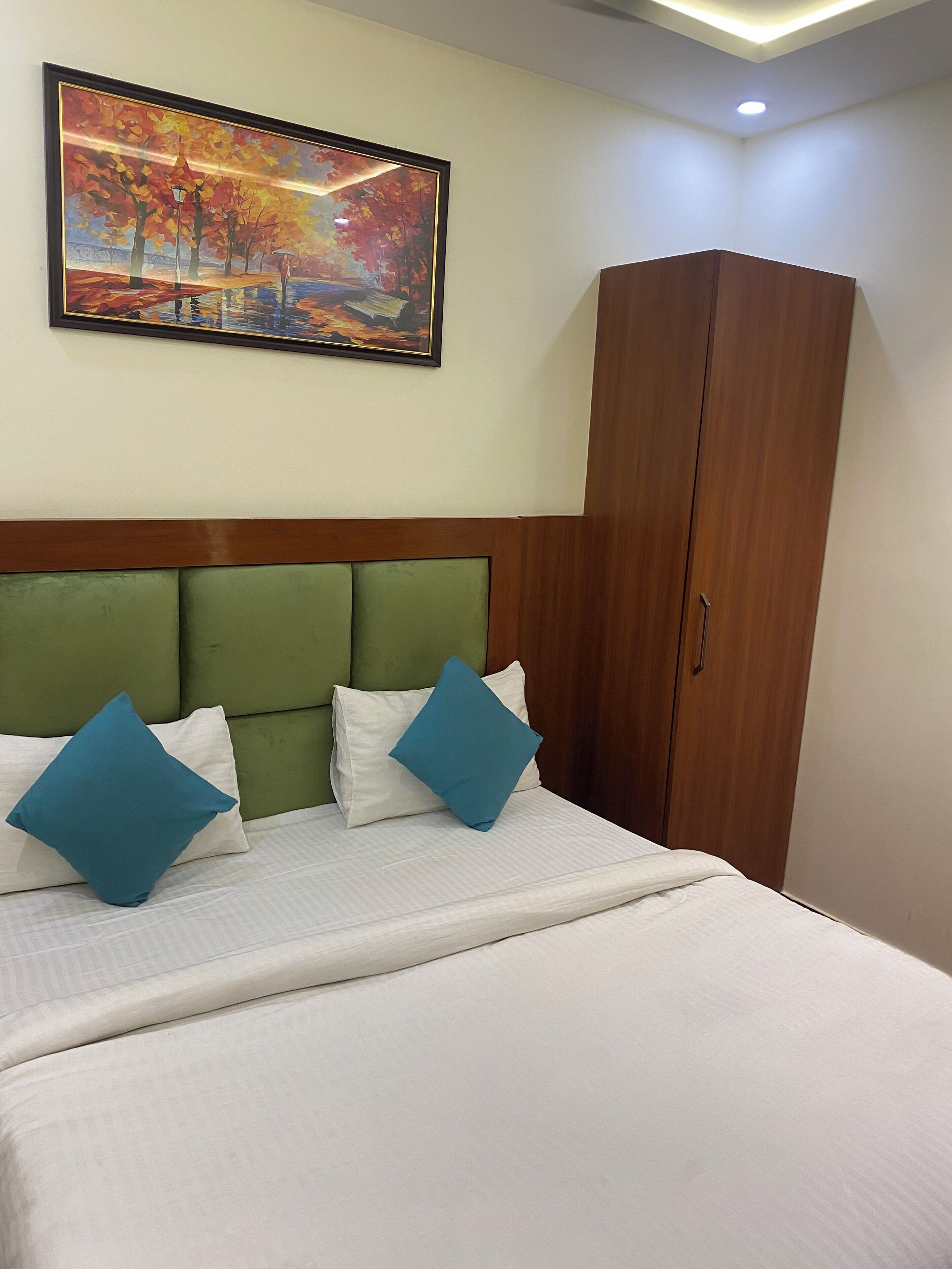 A hotel room with a bed, green headboard, white pillows, a blue pillow, a wooden wardrobe, and a painting of autumn trees on the wall.
