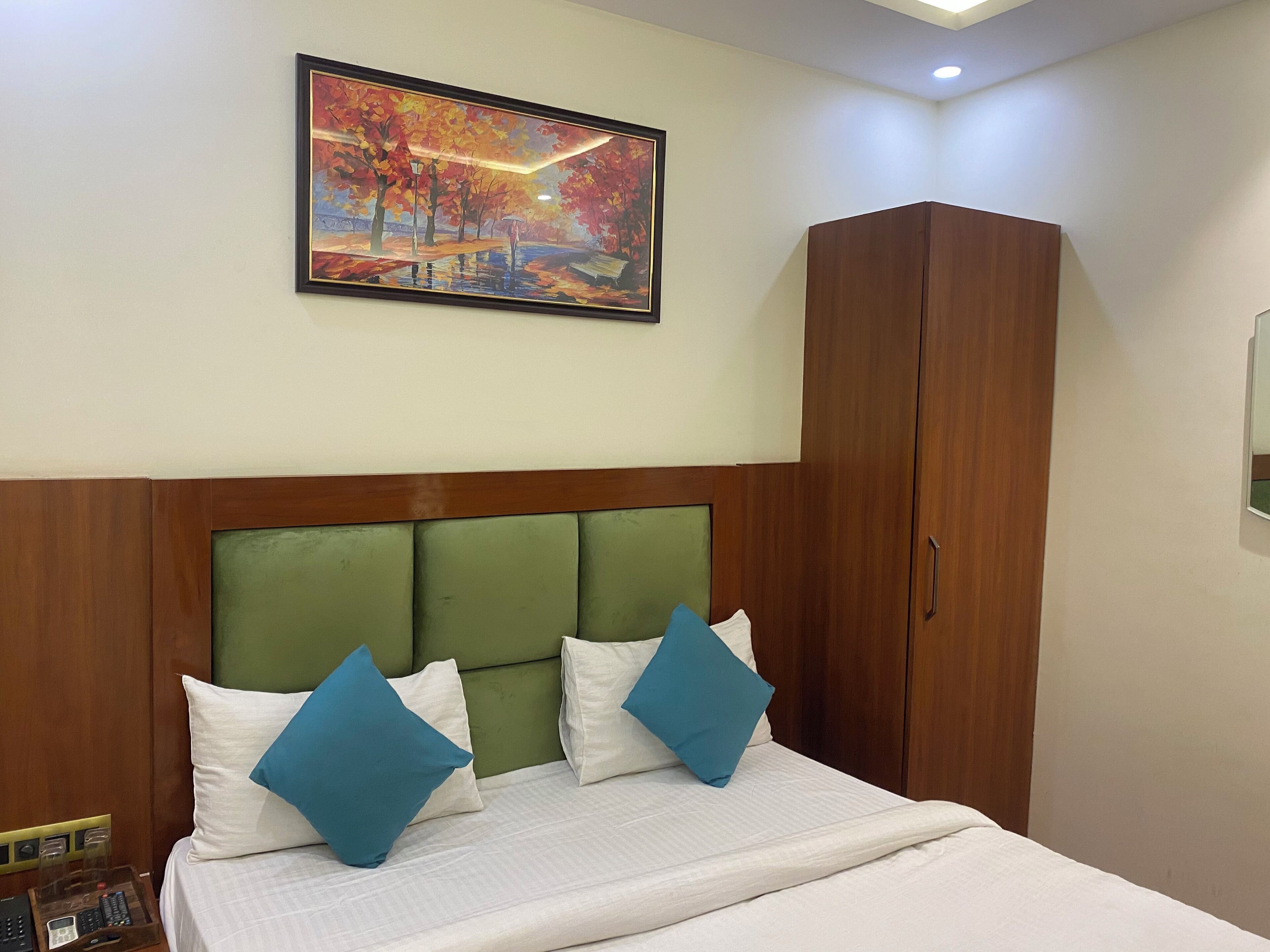 Habitación de hotel con cama, cabecera verde, ropa de cama blanca, almohadas azules, un armario de madera y un cuadro enmarcado con un parque.