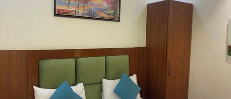 Habitación de hotel con cama, cabecera verde, ropa de cama blanca, almohadas azules, un armario de madera y un cuadro enmarcado con un parque.