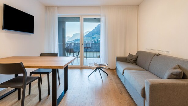 Ein modernes Wohnzimmer mit Holztisch, schwarzen Stühlen, einem grauen Sofa, einem Flachbildfernseher und einer Glastür, die auf einen Balkon mit Bergblick führt.